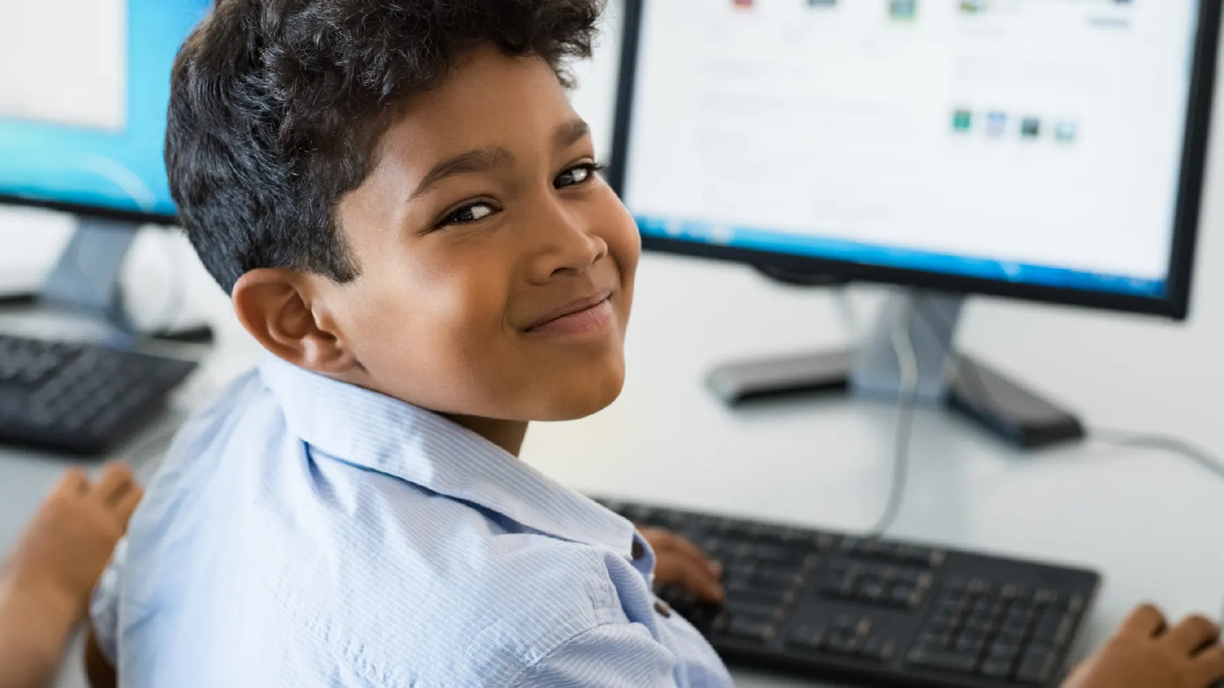 An engaged and happy school aged boy seated at a computer looking at the camera over his shoulder