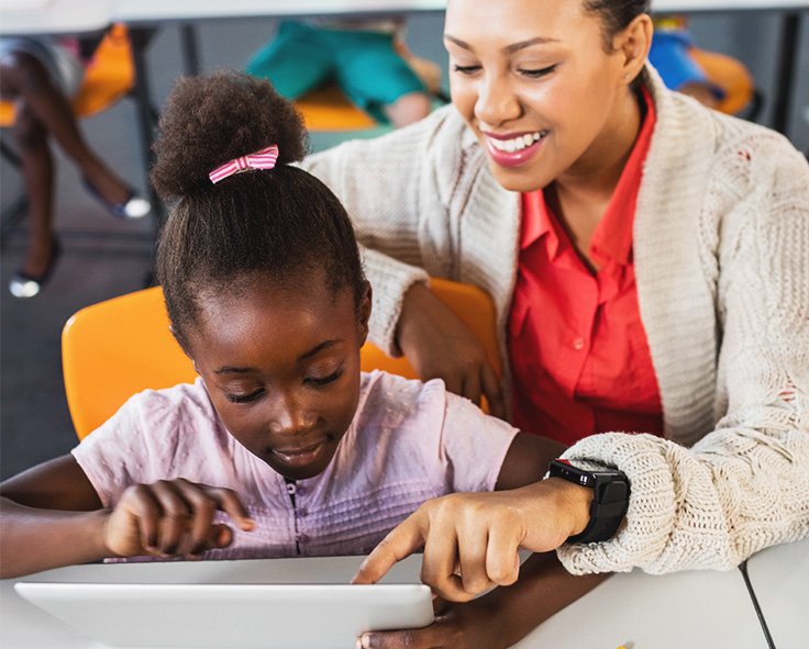A teacher in a classroom guiding a young child through a classroom resource on a tablet device