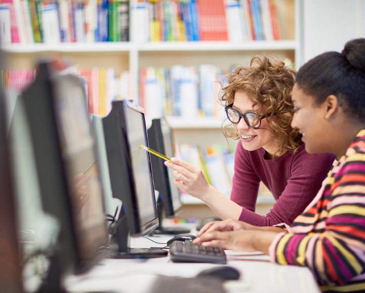 A teacher pointing out a detail in a students coursework to the student using Snapplify on a desktop computer in a library