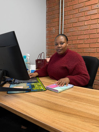 Mapule-in-office - Snapplify Education Woman sits at a wooden desk in an office, facing the camera, wearing a burgundy sweater with a computer monitor and notebook on the desk.